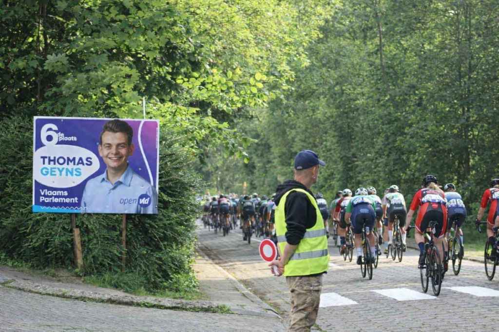 Un groupe de cyclistes traverse Tervuren, avec à gauche une affiche électorale de Thomas Geyns (Voor Tervuren, Anders/Open VLD).